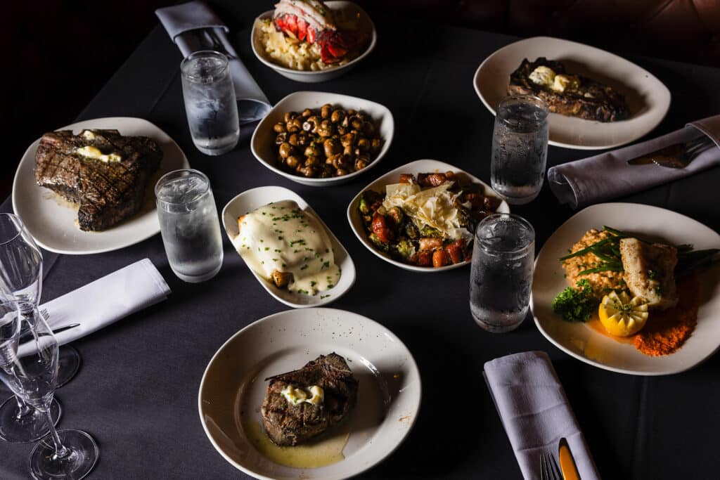 A table full of Bowdie's top-selling steaks and sides including Lobster Mac, Au Gratin Potatoes, and Mushrooms.